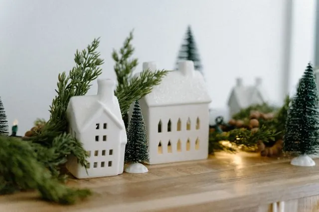 Small white ceramic Christmas houses with fir sprigs and bottle brush trees on a wooden shelf