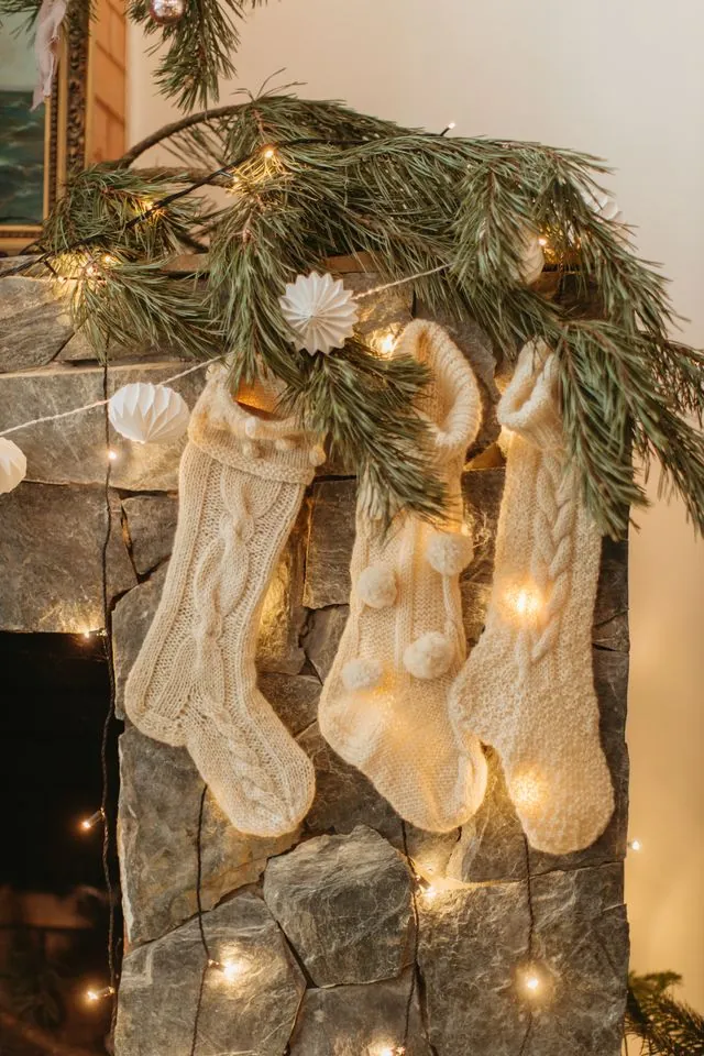 Cream knit Christmas stockings hanging from a stone fireplace with a lit garland