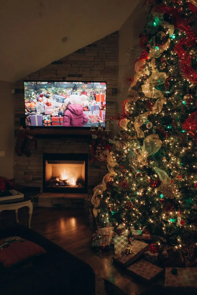 Christmas mantel kept low under a TV above a lit fireplace, next to a decorated tree