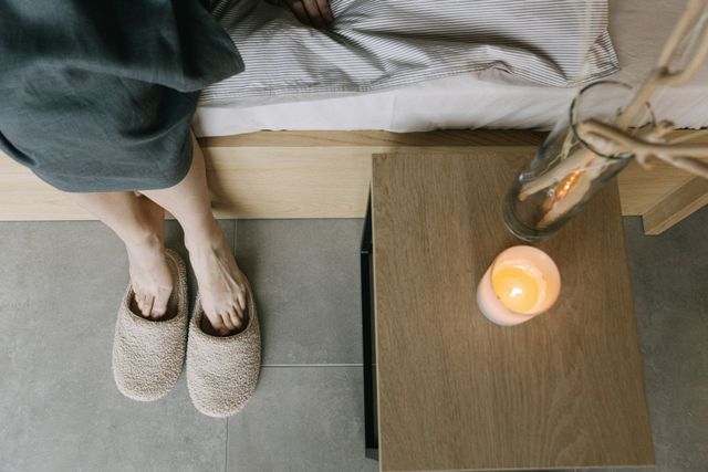Woman wearing soft felt house slippers next to a bedside table with a lit candle