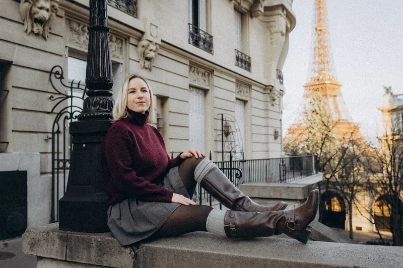 Woman in a burgundy turtleneck sweater, plaid skirt and brown boots sitting on a stone ledge in Paris, one of the sweater outfits in this post