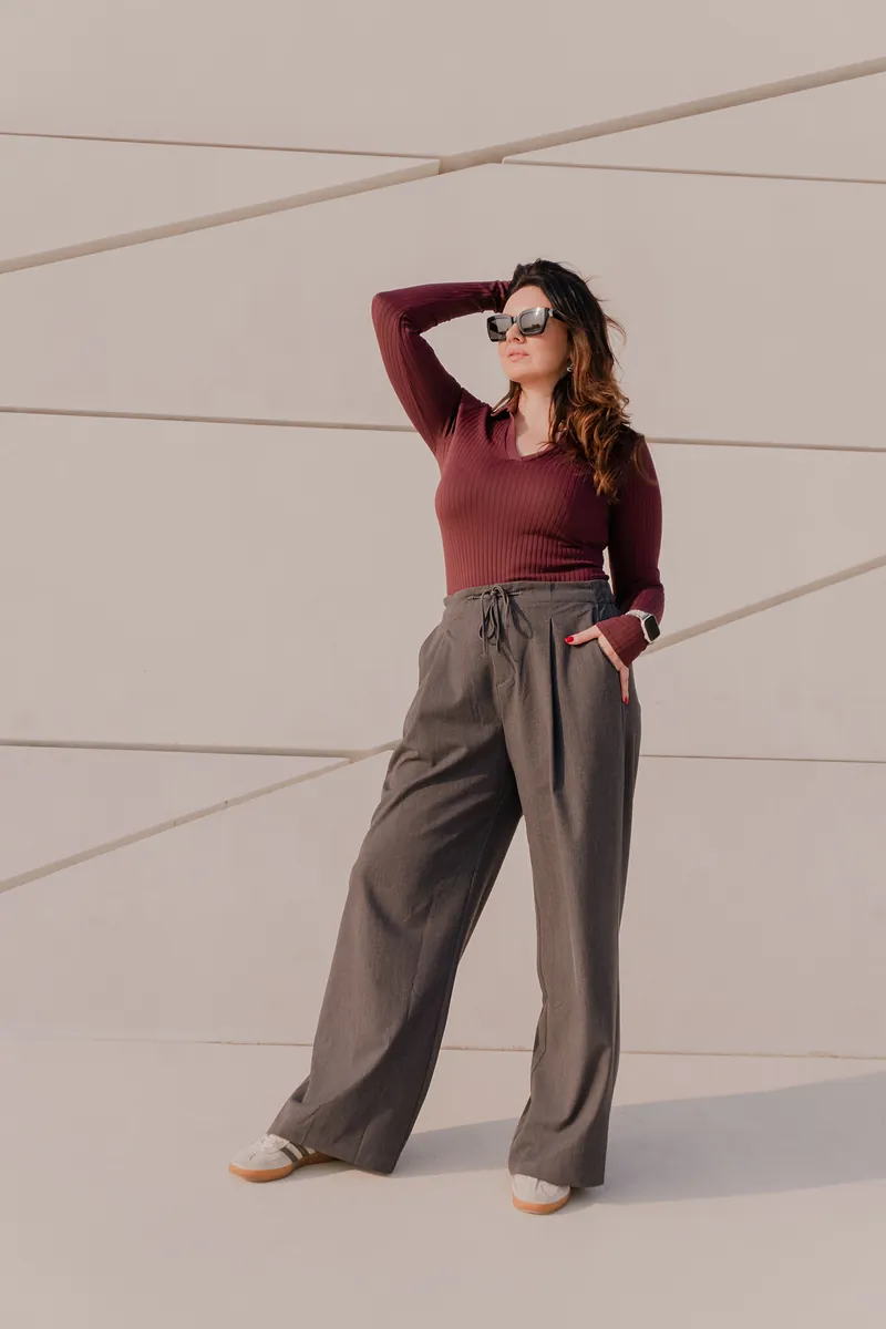 Woman in a burgundy top and wide brown trousers standing against a pale wall, a sweater outfit with brown trousers